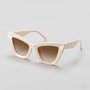 May include: Cat-eye sunglasses featuring a thick, cream-coloured plastic frame with sharp, angular edges. The lenses are gradient, transitioning from a darker brown at the top to a lighter, translucent brown at the bottom, set within the frame.