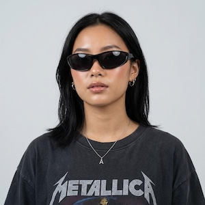 May include: Oval-shaped black sunglasses. The person in the image is wearing a black t-shirt with the word "METALLICA" printed on it. They also have a silver necklace and hoop earrings. The background is plain white.