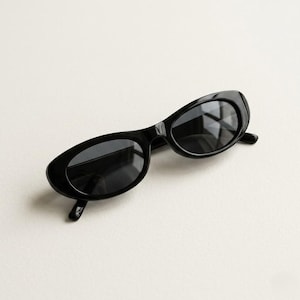 May include: A pair of black oval sunglasses with dark lenses. The sunglasses feature a thick, glossy black frame and a retro design. The dark lenses offer protection from the sun. A fashionable accessory for any look, these sunglasses are a classic style.