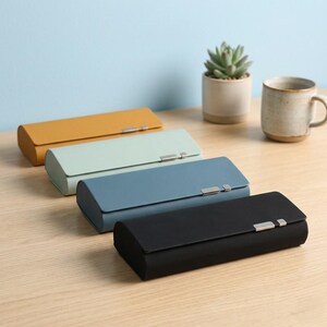 May include: Four rectangular eyeglass cases arranged in a diagonal row. The cases are mustard yellow, light mint green, dusty blue, and black. Each case features a smooth, solid-colored exterior with a small, rectangular silver-toned metal clasp.
