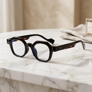 May include: Black eyeglasses with a geometric frame and tortoise-shell arms. The glasses are set on a white marble surface. The lenses are clear.