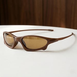 Puede incluir: Gafas de sol marrones con un diseño de montura envolvente y un acabado liso y brillante. Las lentes están tintadas en un tono marrón a juego, lo que proporciona un aspecto coherente. Las patillas son finas y se extienden desde los bordes.