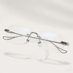 May include: Rimless eyeglasses with a silver metal frame. The temples and bridge feature decorative embellishments. The lenses are rectangular and clear. The glasses are displayed on a white surface.