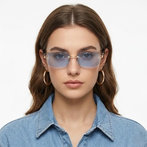 May include: Rimless sunglasses with gold-colored frames and light blue lenses. The rectangular lenses are set in a gold-colored metal frame. The sunglasses are worn by a person wearing a denim shirt and gold hoop earrings.