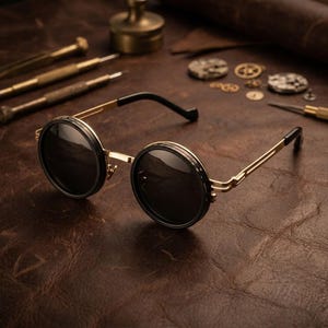 Puede incluir: Gafas de sol redondas con lentes negras y montura dorada. Las gafas tienen un borde negro alrededor de las lentes y patillas negras. Las gafas están sobre una superficie de cuero marrón, con herramientas de relojería en el fondo.