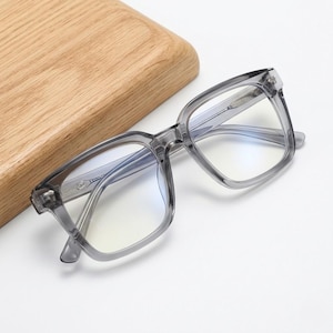 Könnte beinhalten: Graue Brille mit quadratischem Rahmen und klaren Gläsern. Die Brille liegt auf einer weißen Oberfläche, im Hintergrund ein Holzschneidebrett. Die Rahmen haben ein subtiles, durchscheinendes Aussehen.