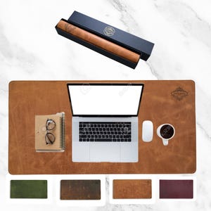 May include: A rectangular tan leather desk mat featuring a laptop, a notebook with glasses, a computer mouse, and a coffee mug. A matching tan leather roll is inside a dark blue gift box. Four additional desk mats in green, brown, tan, and maroon.