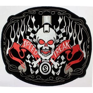 May include: An oval-shaped embroidered patch featuring a central white skull with red eye sockets and teeth. A red banner across the skull reads "SPEED FREAK" in white letters. The design includes silver flames, checkered patterns, and a letter S.