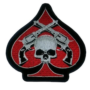 May include: An embroidered patch in the shape of a spade suit symbol. The background is red with a textured pattern, bordered in black. Centered is a silver skull with two crossed silver revolvers positioned behind it, all outlined in black.