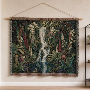 May include: A rectangular woven wall tapestry featuring a detailed landscape scene of a cascading waterfall surrounded by lush green foliage, ferns, and various colourful flowers. The tapestry has a fringed border and is suspended from a wooden rod.