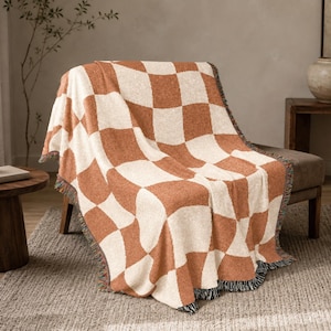 May include: A woven throw blanket featuring a large-scale checkered pattern in alternating terracotta orange and cream white squares. The blanket has a textured, knit-like appearance with frayed, multi-colored fringe edges along the bottom border.