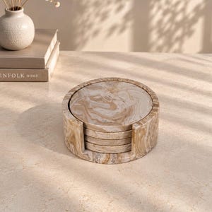 May include: A set of circular coasters featuring a swirling beige and white marbled pattern, stacked neatly within a matching cylindrical holder. The coasters and the holder display a consistent, polished finish with organic, flowing colour veins.