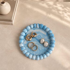 May include: A round blue and white marbled tray with a raised, beaded edge. The tray holds two gold-toned hoop earrings, two gold-toned rings, and a pair of oval-shaped white pearl-like earrings resting on the smooth, swirling surface.