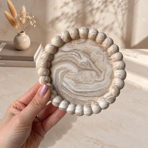 May include: A circular tray featuring a swirling beige, cream, and white marbled pattern on its flat surface. The outer edge is decorated with a continuous row of small, rounded spheres that match the marbled color scheme of the center.
