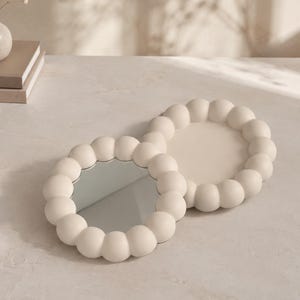 May include: Two circular objects with a matte off-white finish and a raised, bubble-like border. One object is a mirror with a reflective glass center, while the other is a tray with a solid, flat, off-white circular base.