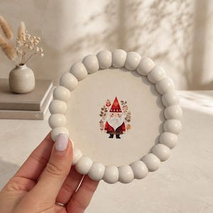 May include: A circular coaster featuring a white beaded frame surrounding a central illustration of a gnome wearing a red hat and dark clothing, accented by botanical elements in shades of red, orange, and brown on a cream-colored background.