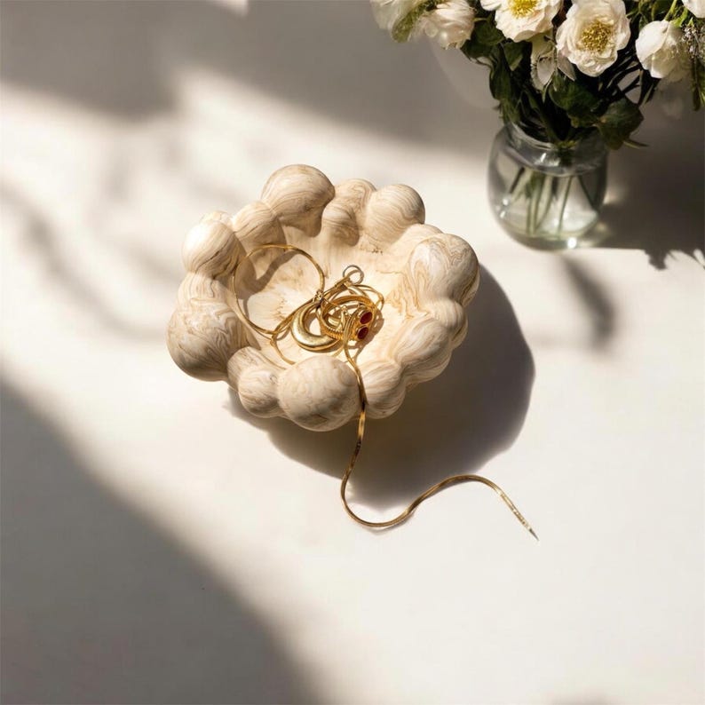 May include: A beige, marbled, cloud-shaped trinket dish with a bumpy, rounded rim. Inside the dish rests a collection of gold-toned jewellery, including a thin chain necklace and several rings, one of which features a small, dark red stone accent.