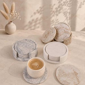 May include: A collection of circular stone coasters featuring marbled patterns in shades of grey, beige, and cream. The set includes two cylindrical holders and a matching cup resting on a grey marbled coaster, all arranged on a neutral surface.