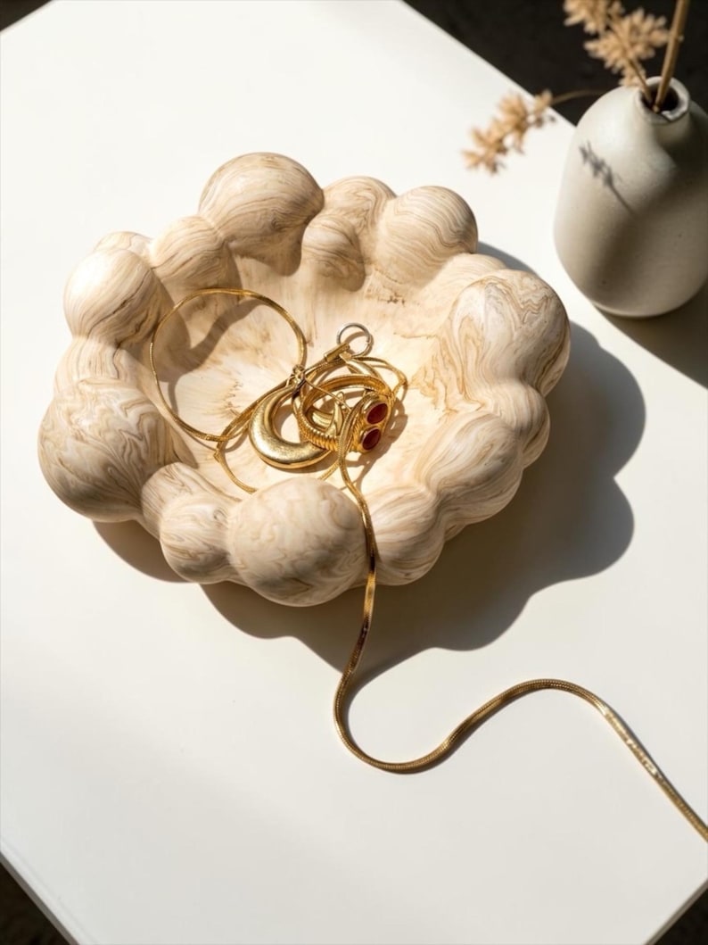 May include: A beige and cream marbled bowl with a rounded, bubble-like exterior texture. Inside the bowl sits a collection of gold-toned jewellery, including a thin chain necklace, a circular bangle, and a ring with two small red stones.