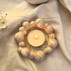 May include: A circular candle holder with a marbled, swirled pattern in shades of cream, beige, and soft peach. The holder features a cluster of rounded, bubble-like spheres around the perimeter, containing a single lit white tea light candle.