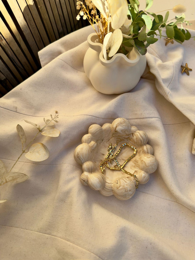 May include: A cream-coloured, cloud-shaped trinket dish with a gold necklace inside. A small, white vase with dried flowers and greenery is nearby. The scene is set on cream-coloured fabric.