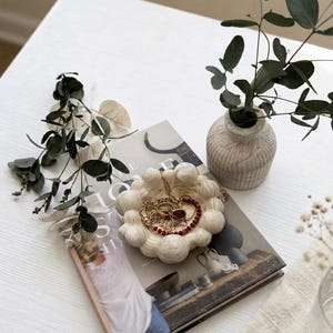 May include: A round, off-white, textured trinket dish with a scalloped edge, containing gold-toned jewellery and a red beaded necklace. The dish rests on a hardcover book titled "The Home Story," accompanied by a small, ribbed, light-coloured vase.