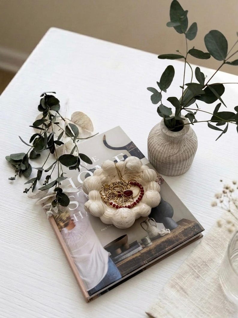 May include: A round, off-white, textured trinket dish with a scalloped edge, containing gold-toned jewellery and a red beaded necklace. The dish rests on a hardcover book titled "The Home Story," accompanied by a small, ribbed, light-coloured vase.