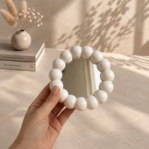 May include: A small, circular mirror featuring a decorative frame composed of numerous smooth, matte white spheres arranged in a ring. The reflective glass center is surrounded by this bubble-like border, creating a distinct, tactile appearance.