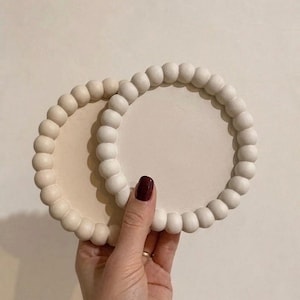 May include: Two round, off-white coasters with a beaded edge. The coasters are held in a hand with a dark red nail polish. The coasters have a smooth, light-colored center.