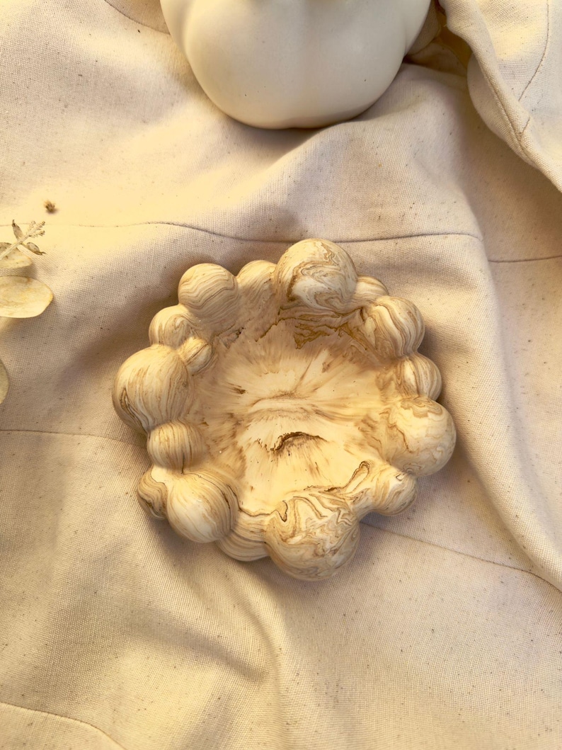 May include: A small, circular decorative bowl featuring a marbled pattern of cream, beige, and brown swirls. The rim is composed of numerous rounded, bubble-like protrusions that create a textured, organic edge around the central basin.