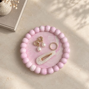 May include: A round tray with a pink and white marbled surface and a border of pink spherical beads. Resting on the tray are two gold hoop earrings with white pearl drops, a thin gold ring, and a white hair clip with a speckled pattern.