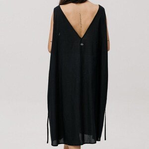 May include: A sleeveless black dress featuring a deep V-neckline on the back, a loose-fitting silhouette, and thin side ties. The garment is made of a textured fabric and includes a small, circular silver-toned metal emblem positioned below the neckline.