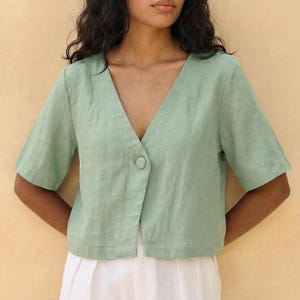 May include: A pale green linen top with a V-neck and a single button fastening. The top has short sleeves and is worn with white trousers. The outfit is simple and elegant, suitable for casual wear.