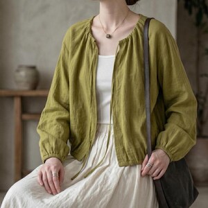 May include: An olive green long-sleeved jacket with a gathered round neckline and thin tie closures. The jacket is worn over a plain white top and a long cream-colored skirt. A dark brown leather shoulder bag strap hangs over the shoulder.