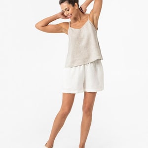 May include: A beige linen camisole top with thin spaghetti straps and a V-neckline paired with white linen shorts featuring an elastic waistband and a relaxed fit, completed by a pair of cream-colored slip-on espadrilles with woven jute soles.