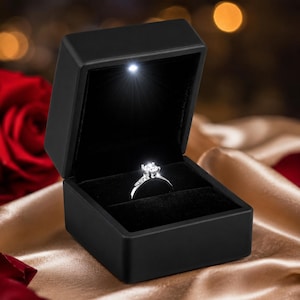 May include: A silver-toned ring with a single clear faceted stone set in a black square jewelry box. The box features an integrated LED light at the top interior, which illuminates the ring from above, highlighting the stone's reflective facets.