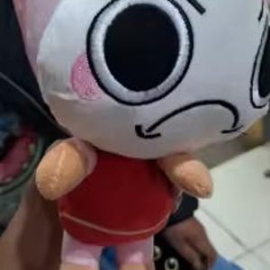Peut inclure: Une peluche avec une t&ecirc;te ronde blanche, de grands yeux noirs, une bouche noire courb&eacute;e et des joues roses. Le corps est rouge avec des bras et des jambes beige clair, et une structure rose en forme d'aileron sur le dessus, rappelant une crevette.