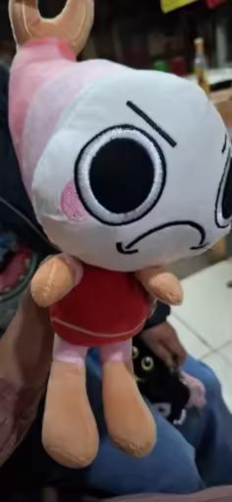 Peut inclure: Une peluche avec une t&ecirc;te ronde blanche, de grands yeux noirs, une bouche noire courb&eacute;e et des joues roses. Le corps est rouge avec des bras et des jambes beige clair, et une structure rose en forme d'aileron sur le dessus, rappelant une crevette.