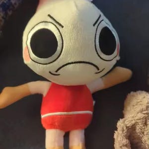 Peut inclure: Une peluche avec une t&ecirc;te ronde couleur cr&egrave;me, de grands yeux noirs, une bouche fronc&eacute;e et des joues roses. La figurine poss&egrave;de une queue de crevette sur le dessus de la t&ecirc;te, porte un haut rouge avec une rayure blanche et a des membres beiges.