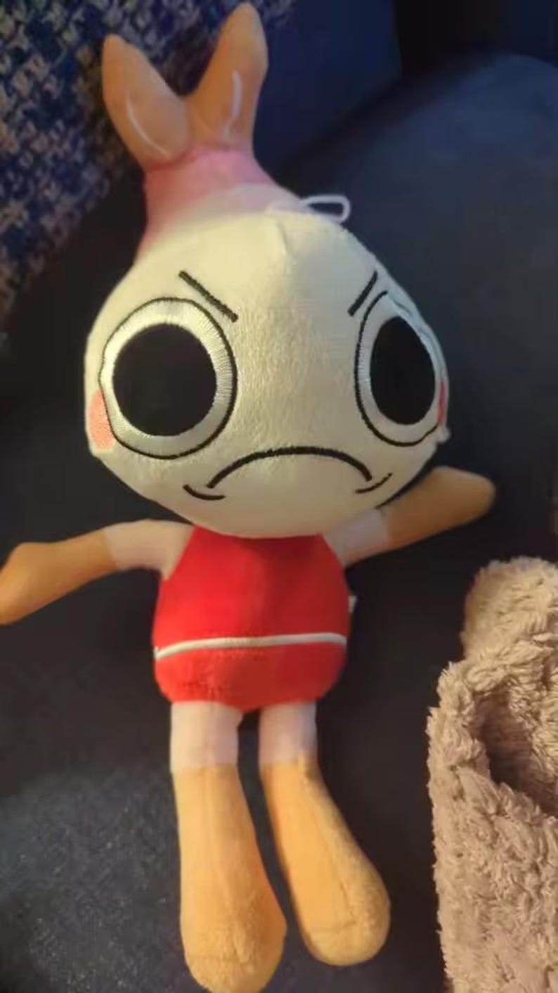 Peut inclure: Une peluche avec une t&ecirc;te ronde couleur cr&egrave;me, de grands yeux noirs, une bouche fronc&eacute;e et des joues roses. La figurine poss&egrave;de une queue de crevette sur le dessus de la t&ecirc;te, porte un haut rouge avec une rayure blanche et a des membres beiges.