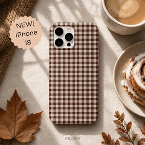 以下が含まれることがあります： ブラウンとクリーム色のギンガムチェック柄の長方形のスマートフォンケース。ケースには3眼カメラシステム用の切り欠きがあります。左側の円形のバッジには「NEW! iPhone 18」というテキストが表示されています。下部にはブランド名「KELINA」が印刷されています。