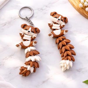 Op de afbeelding: Twee handgemaakte sleutelhangers met een bruin en wit corgi-hondenontwerp. De corgi ligt met zijn poten omhoog, vastgemaakt aan een bruine en witte staart. De sleutelhangers liggen op een marmeren oppervlak.