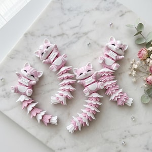 Op de afbeelding: Een verzameling roze en witte katvormige beeldjes met lange, gelaagde staarten. De katten bevinden zich in verschillende speelse poses, met gedetailleerde poten en gezichten. De beeldjes zijn gerangschikt op een wit marmeren oppervlak.
