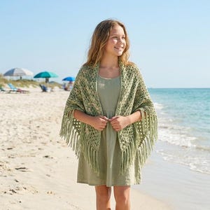 Può includere: Una persona indossa un vestito verde chiaro e uno scialle all'uncinetto con frange, in piedi su una spiaggia sabbiosa. Lo scialle è di colore verde chiaro con un motivo strutturato. Lo sfondo comprende l'oceano, ombrelloni e un cielo azzurro.