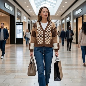 Könnte beinhalten: Eine Frau in einer braunen, cremefarbenen und beigefarbenen karierten Strickjacke und blauen Jeans geht durch ein Einkaufszentrum. Sie trägt eine graue Handtasche und eine schwarze Einkaufstasche. Das Einkaufszentrum hat ein Oberlicht und mehrere Geschäfte.