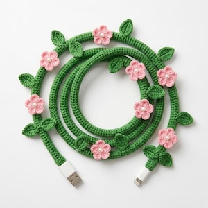May include: A green crocheted charging cable adorned with pink flowers and green leaves. The cable has a USB connector on one end and a lightning connector on the other. The floral design is a decorative accent.