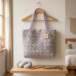 Pode incluir: Uma sacola de crochê feita à mão em tons de lavanda, verde e creme está pendurada em um cabide de madeira. A sacola apresenta um padrão de diamante e uma etiqueta que diz "CROCHET SHRUG HANDMADE". Uma bola de lã, uma agulha de crochê e uma tesoura estão em uma prateleira de madeira.