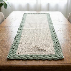 May include: A rectangular, cream-colored crochet table runner with a sage green scalloped border. The runner is displayed on a wooden table, showcasing its texture and design. This handmade piece adds a touch of rustic charm to any dining setting.