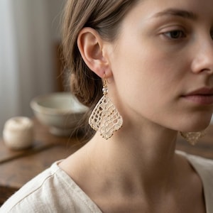 May include: Cream-colored, open-weave lace earrings with gold-colored accents and a small clear bead. The earrings are diamond-shaped and hang from gold-colored hooks. The earrings are a delicate, handmade accessory.