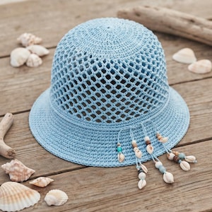 Puede incluir: Un sombrero de pescador de ganchillo azul claro con un tejido abierto de patrón de diamantes en la copa y un ala sólida. El sombrero está decorado con varios hilos colgantes de color azul claro, cada uno con cuentas de madera y conchas.